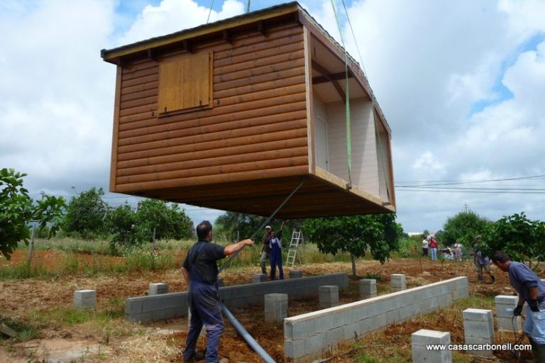 Casa prefabricada en proceso de levantamiento sobre cimientos, rodeada de vegetación.