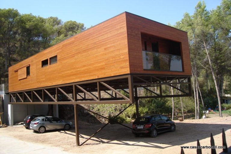 Casa prefabricada moderna de madera elevada sobre estructura metálica, con coches estacionados debajo.