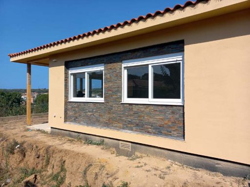 Casa en construcción con paredes de piedra y ventanas modernas, rodeada de terreno.