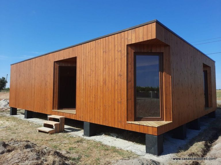 Casa prefabricada moderna de madera con ventanas grandes, ubicada en un terreno despejado. Montaje en España