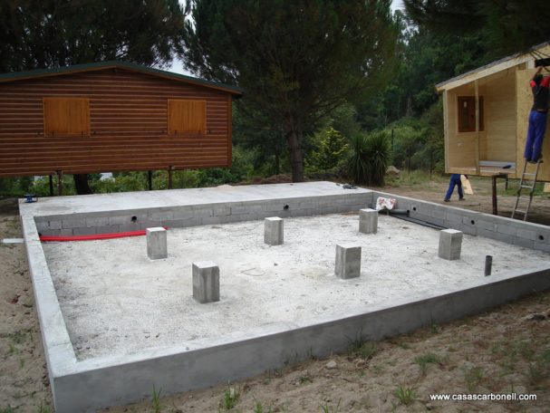 Base de muros corridos y pilares sobre losa de hormigón soterrada Base de concreto con columnas en un terreno, rodeada de árboles y construcciones de madera.