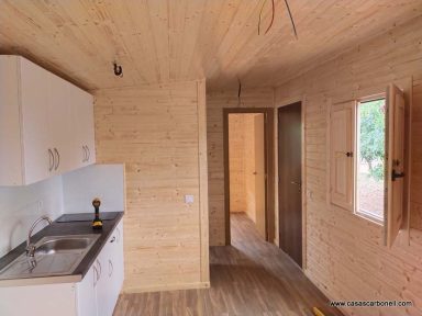 Interior de una cabaña de madera con cocina y puertas, iluminada y en construcción.