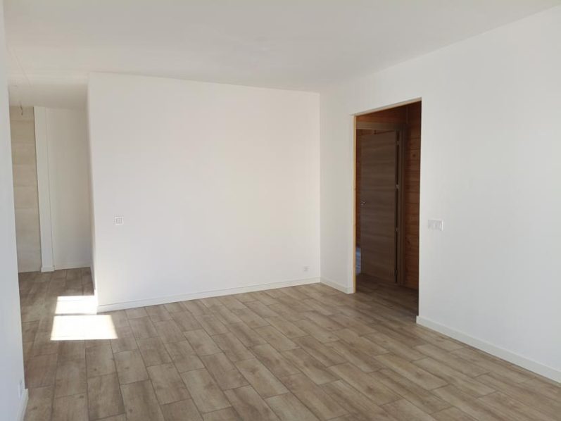 Habitación vacía con paredes blancas y suelo de madera clara, con puerta de madera.