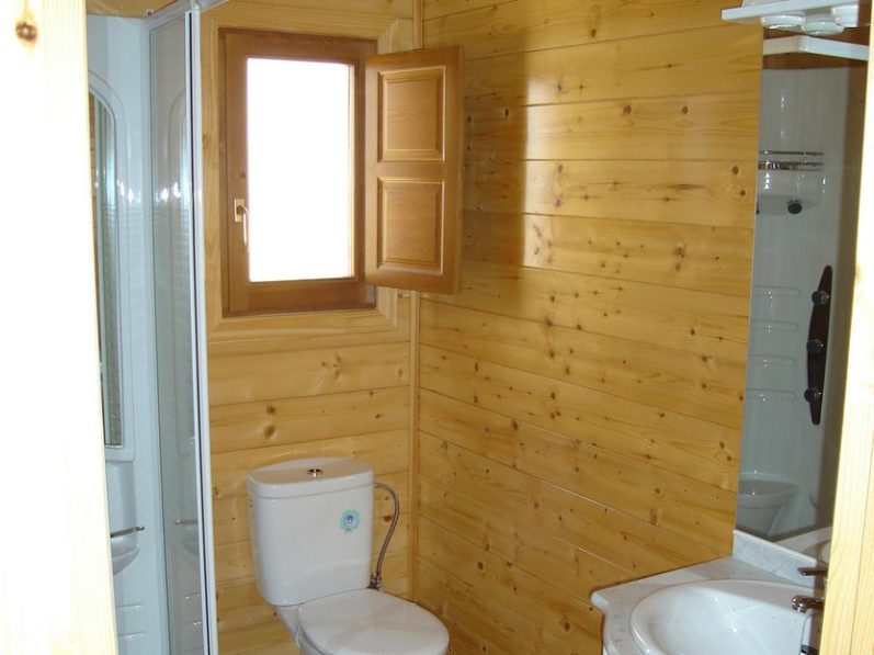 Baño con paredes de madera, inodoro y ducha. Ventana abierta con luz natural.