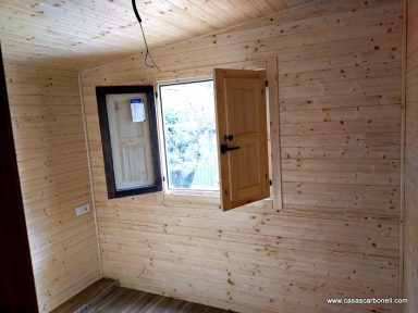 Interior de una habitación de madera con una ventana abierta y una puerta cerrada.